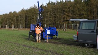 2 Personen stehen vor einer blauen Rammkernsonde auf einem Acker. Kachel Vorschau