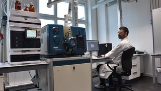 Eine Person sitzt in einem Labor und schaut nach links auf ein großes Analysegerät. Kachel Vorschau