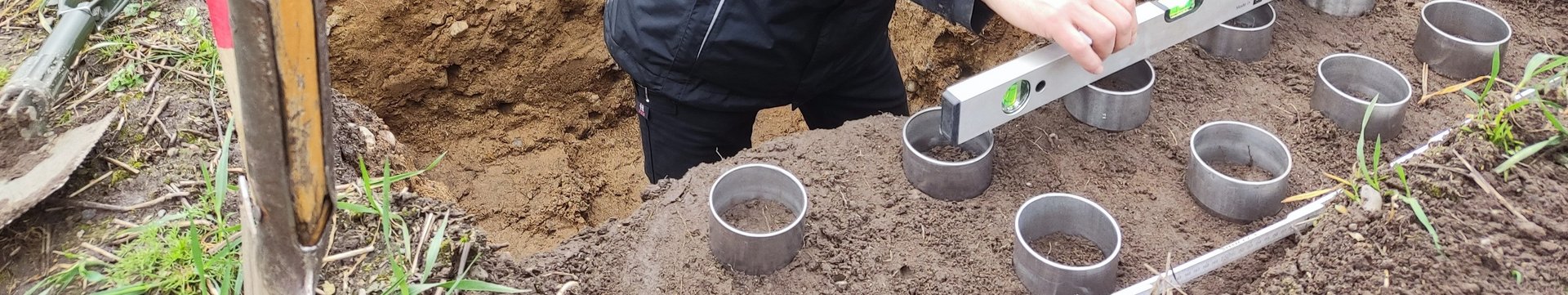 Ein Mann, steht in einer Bodengrube, vor ihm sind Stechzylinder in den Boden geschlagen und er prüft mit einer Wasserwaage die gleichmäßige Einschlagtiefe.