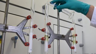 Eine Person im Labor mit weißem Kittel und grünen Handschuhen füllt eine durchsichtige Flüssigkeit in hohe Glassäulen. Kachel Vorschau