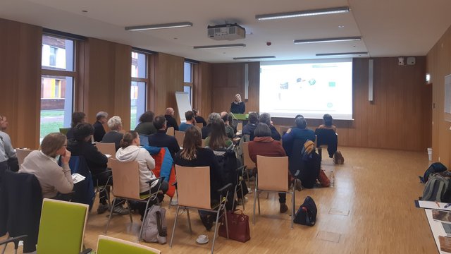 Eine Person hält einen Vortrag mit einer Präsentation vor einem Publikum. Kachel Vorschau