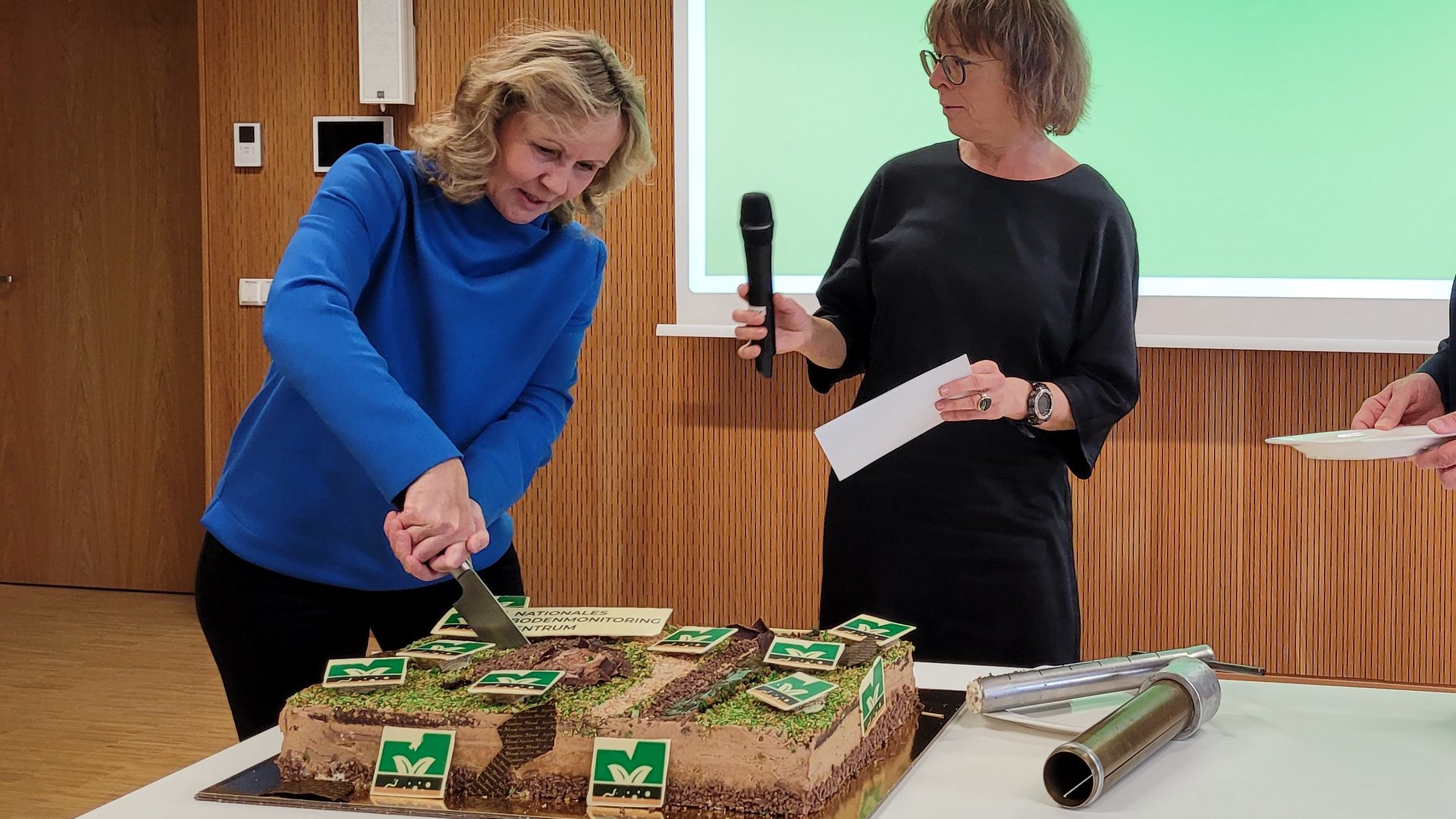 1 Person schneidet eine Torte an, die Moderatorin hält das Mikrofon.