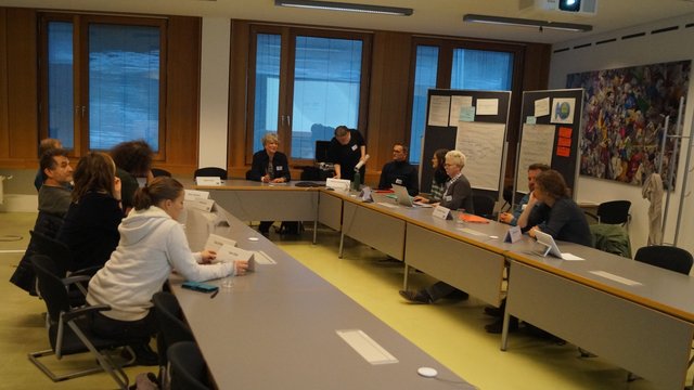 An einem U-förmigen Tisch sitzen viele Personen und diskutieren verschiedene Fragen. Kachel Vorschau