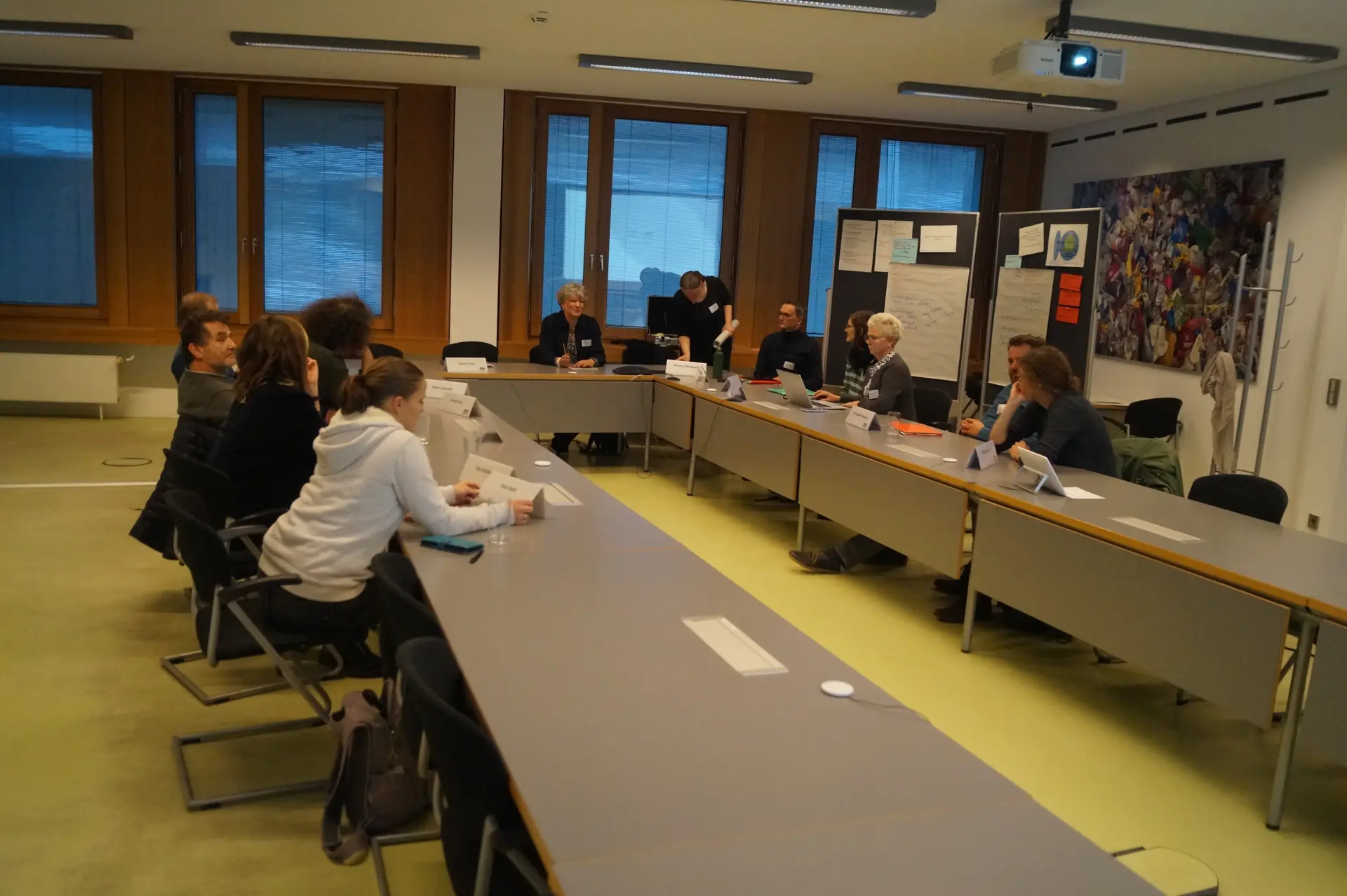 An einem U-förmigen Tisch sitzen viele Personen und diskutieren verschiedene Fragen.