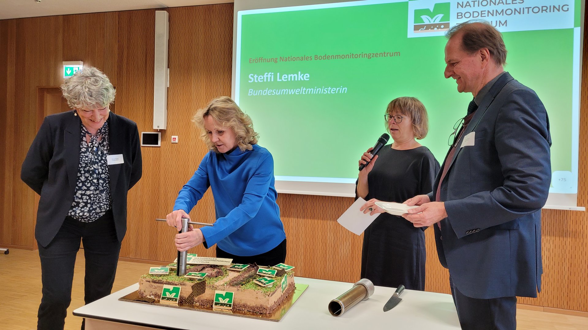 4 Personen stehen um eine Torte, die von einer Person angeschnitten wird.